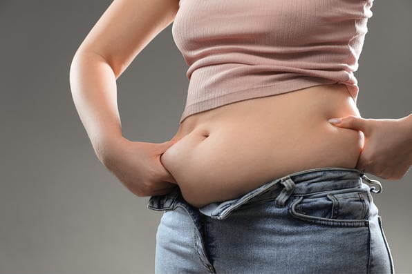 A close-up photo of a person's midsection, showing their stomach and waist area. They are wearing a pink ribbed crop top and low-rise light blue denim jeans. The person is gripping the skin on their sides with both hands. The background is a neutral gray.
